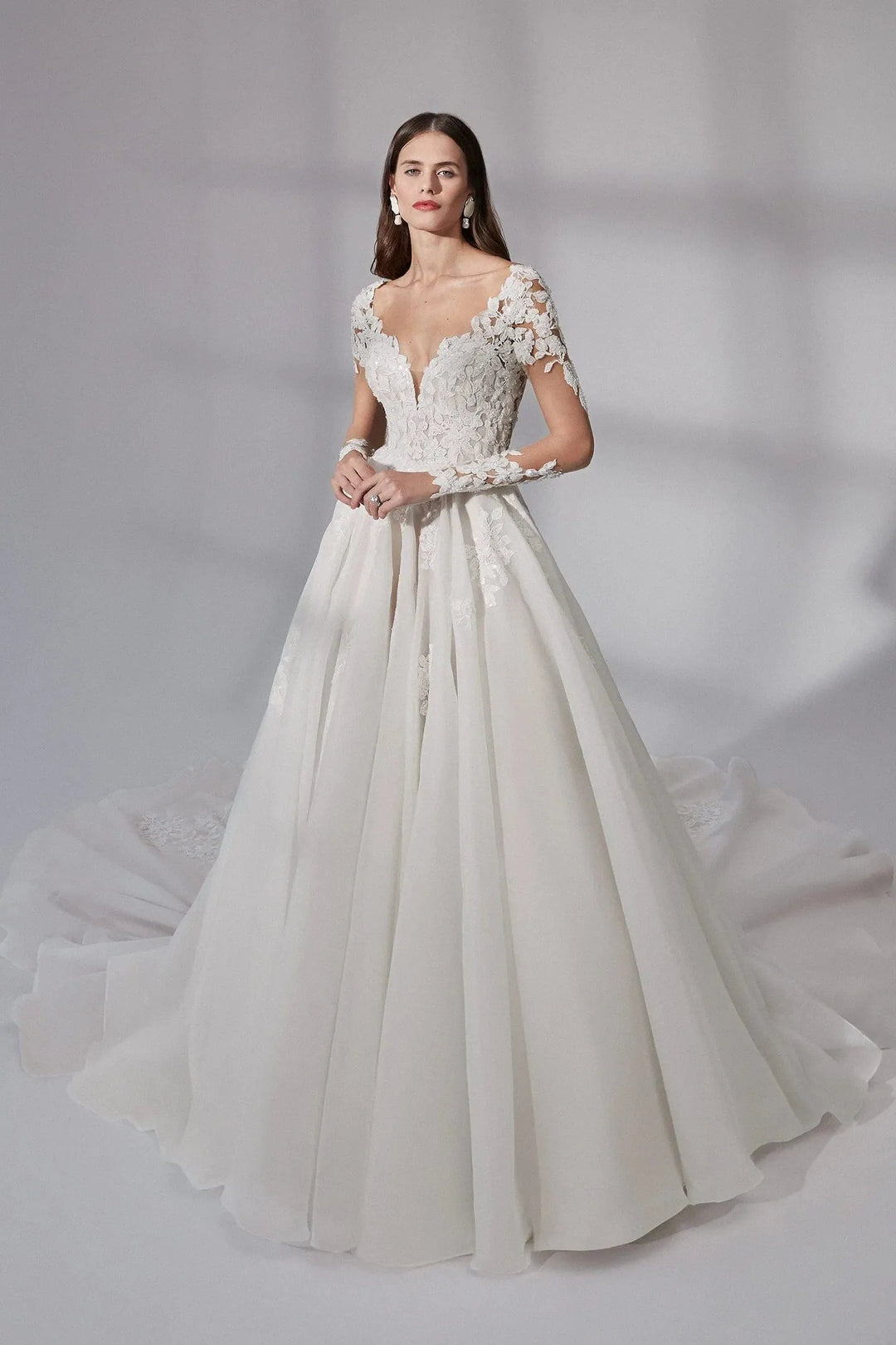 A woman modeling a white ball gown wedding dress with lace bodice, short lace sleeves, a deep V-neckline, and a full flowing skirt against a soft gray studio backdrop.