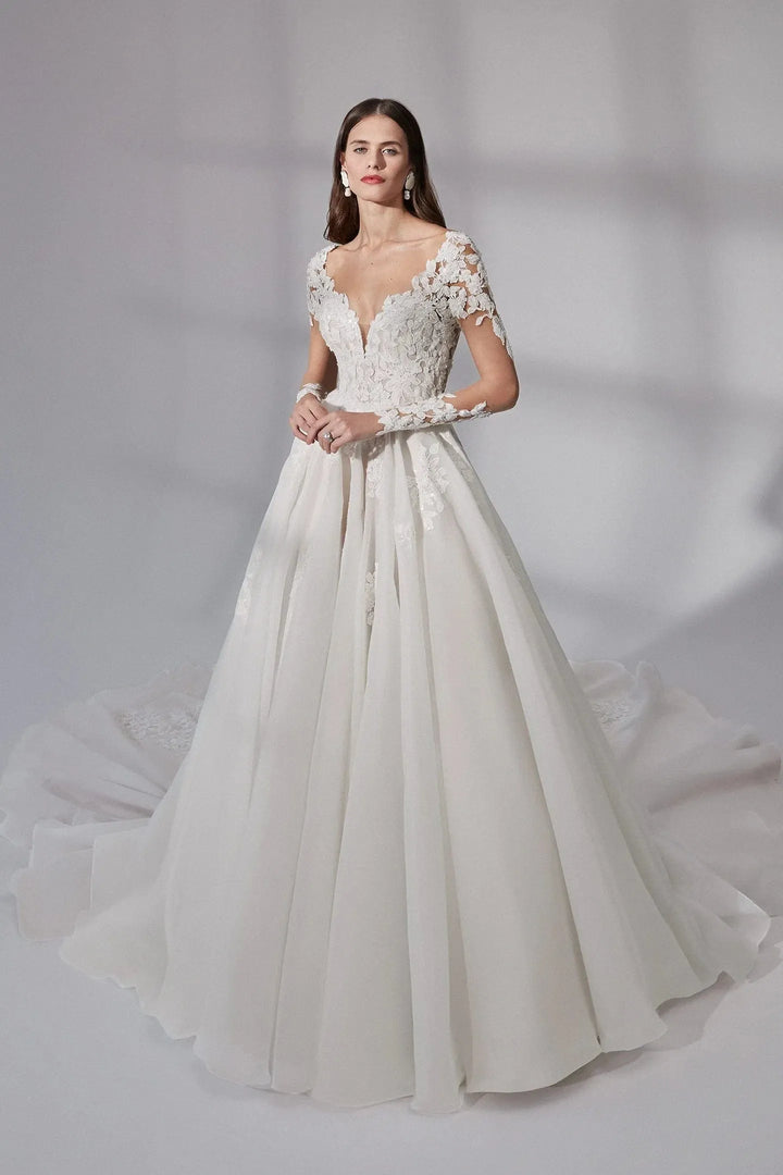 A woman modeling a white ball gown wedding dress with lace bodice, short lace sleeves, a deep V-neckline, and a full flowing skirt against a soft gray studio backdrop.
