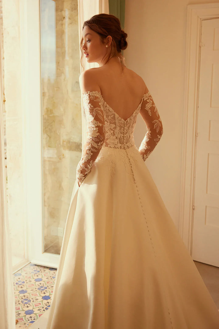 Woman in a white lace wedding dress standing by a window.