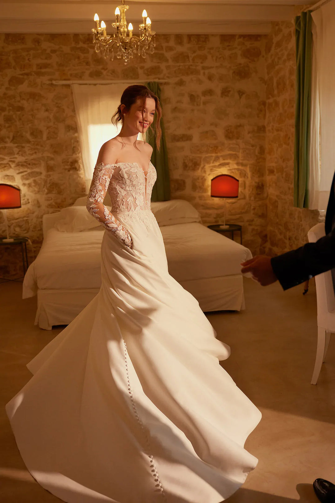 Woman in a white wedding dress standing in a warmly lit bedroom.