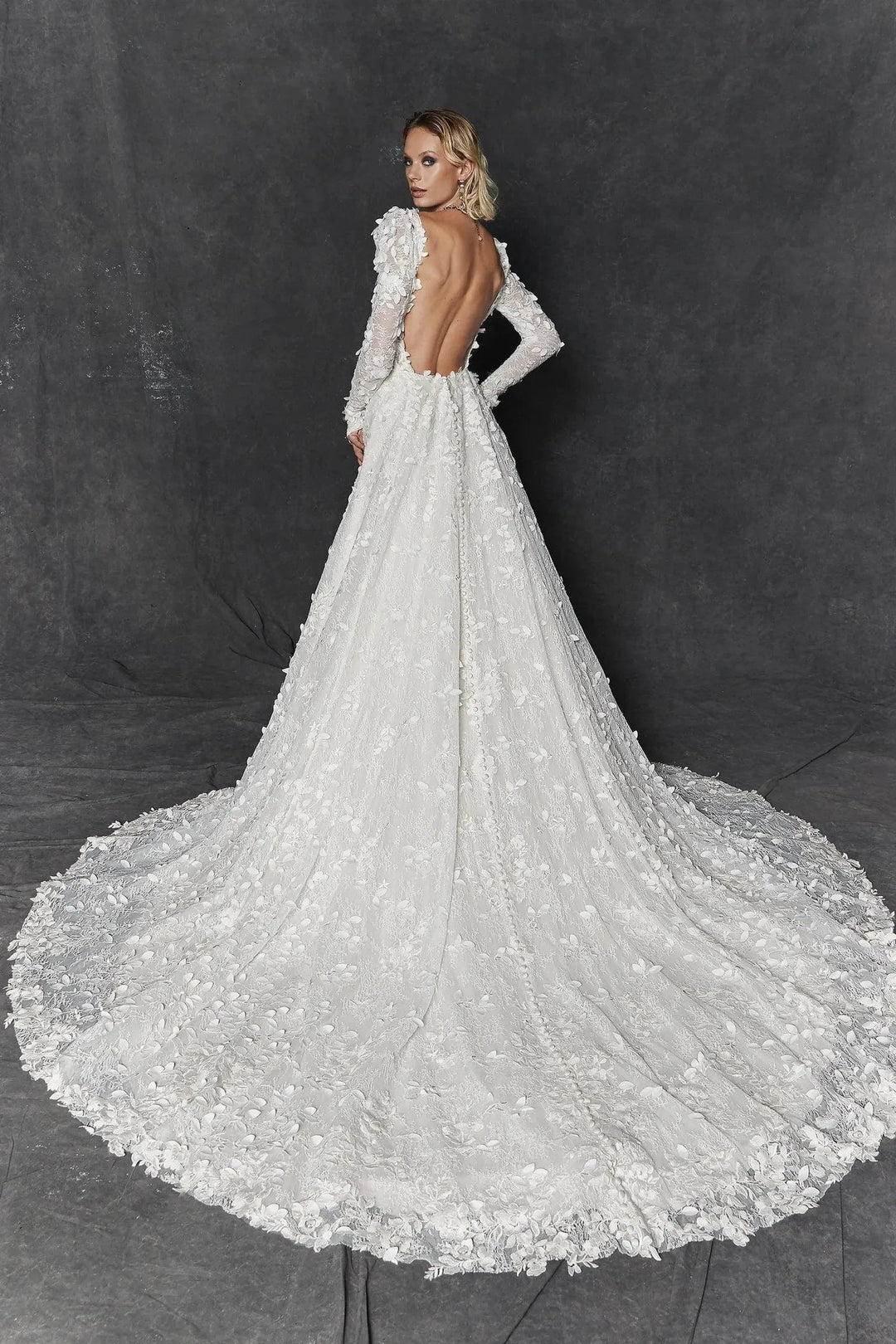 A woman modeling a white bridal gown from the back against a dark gray studio backdrop. The gown has long sleeves, an open back, and a dramatic full train with floral lace and tulle detailing.