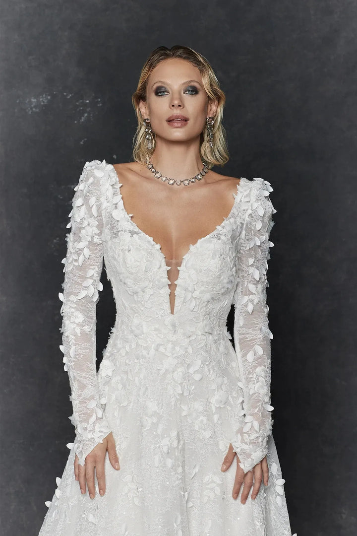 A woman modeling a white long-sleeve wedding dress with 3D floral applique, a deep V-neckline, and lace-up front details, set against a dark textured studio background.