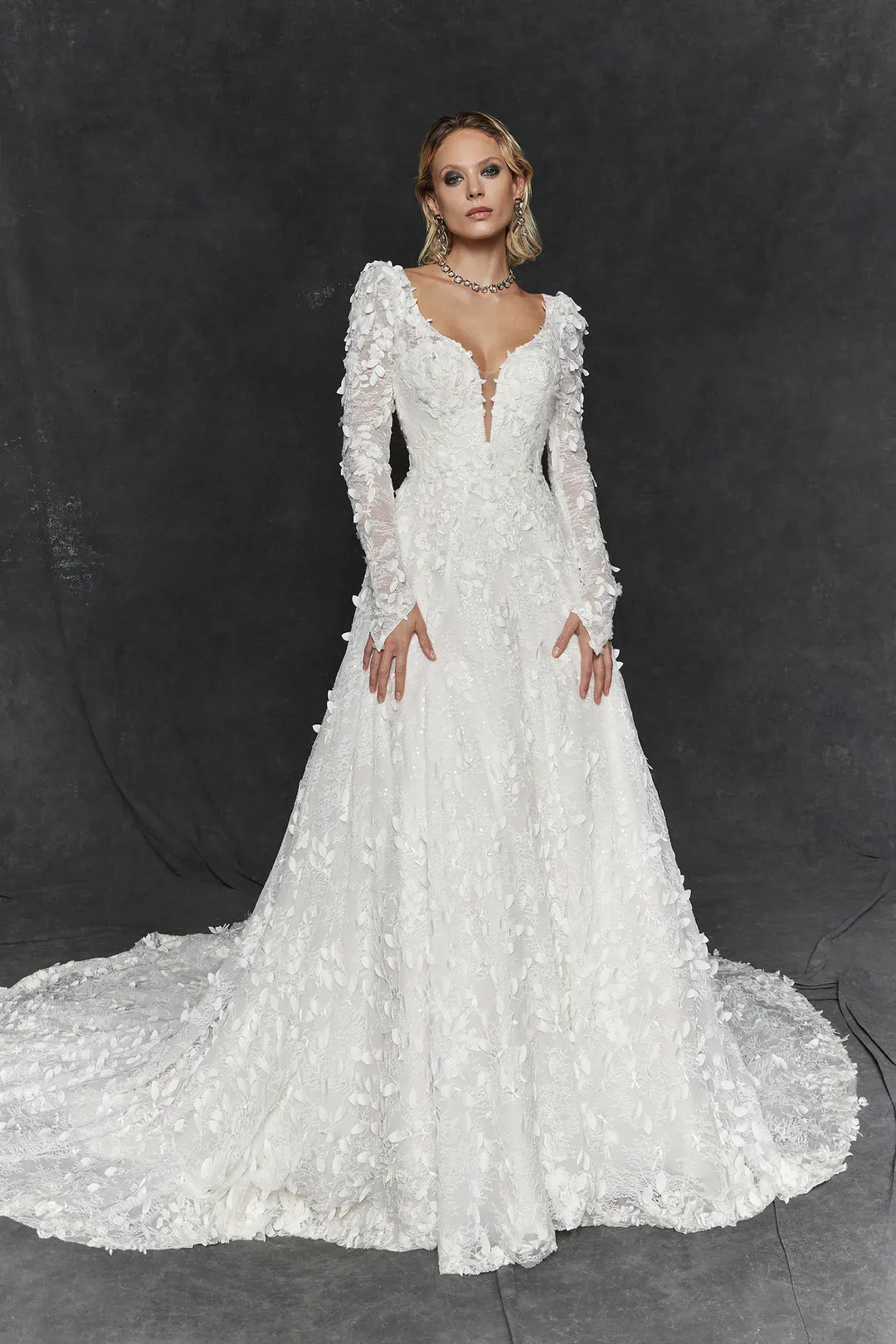 A model wearing a long-sleeved white lace wedding gown with a deep V-neckline and a full skirt with a train, posed against a dark gray studio backdrop.