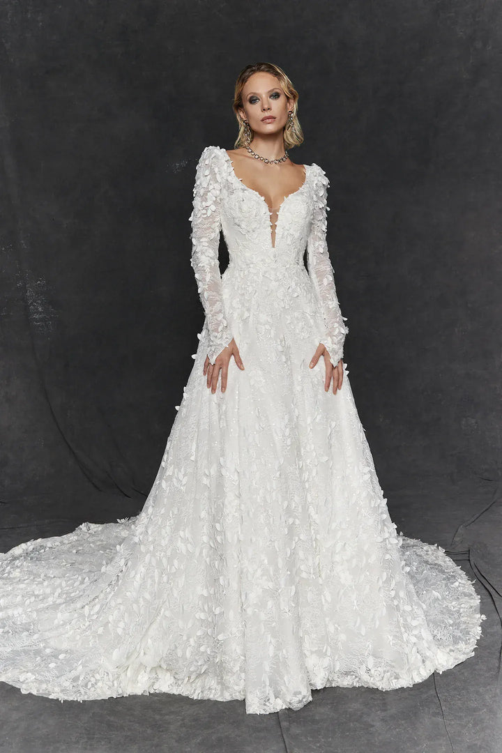 A model wearing a long-sleeved white lace wedding gown with a deep V-neckline and a full skirt with a train, posed against a dark gray studio backdrop.