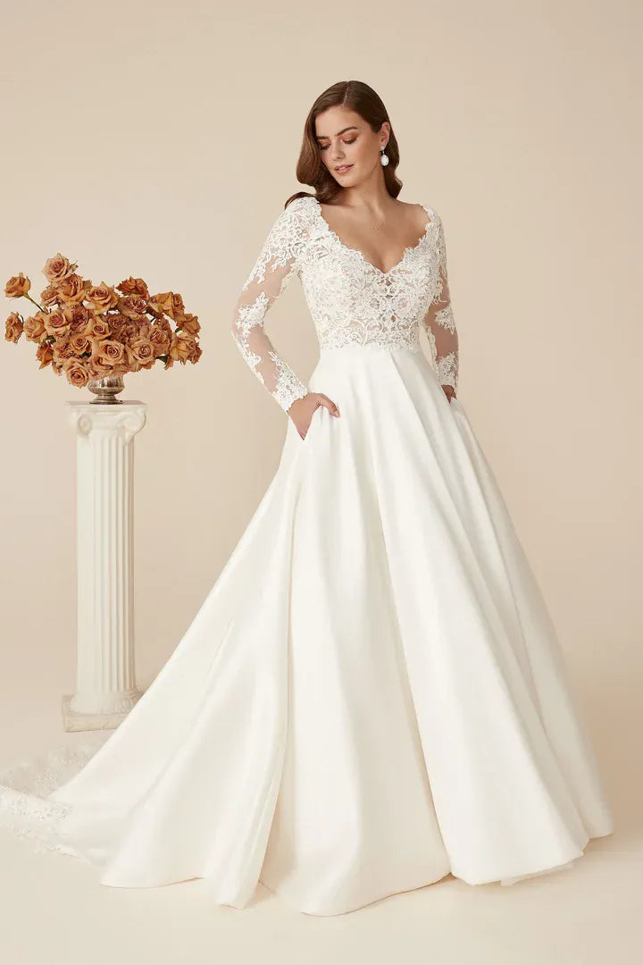 A woman models a white wedding gown with a lace bodice, long sheer sleeves, and a full satin skirt with pockets, posed next to a decorative column with dried flowers against a warm beige studio background.