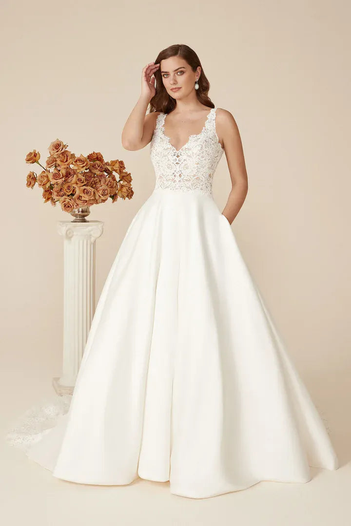 A woman modeling a white wedding gown with a lace-embroidered bodice and a full satin A-line skirt, standing in a studio with a beige backdrop and a decorative pedestal of dried flowers.