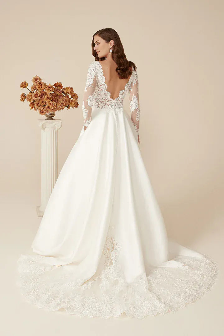 A woman modeling a white wedding gown with long lace sleeves, a deep V back, and a full satin skirt with a lace-trimmed hem, posed in front of a beige studio backdrop beside a pedestal with dried flowers.