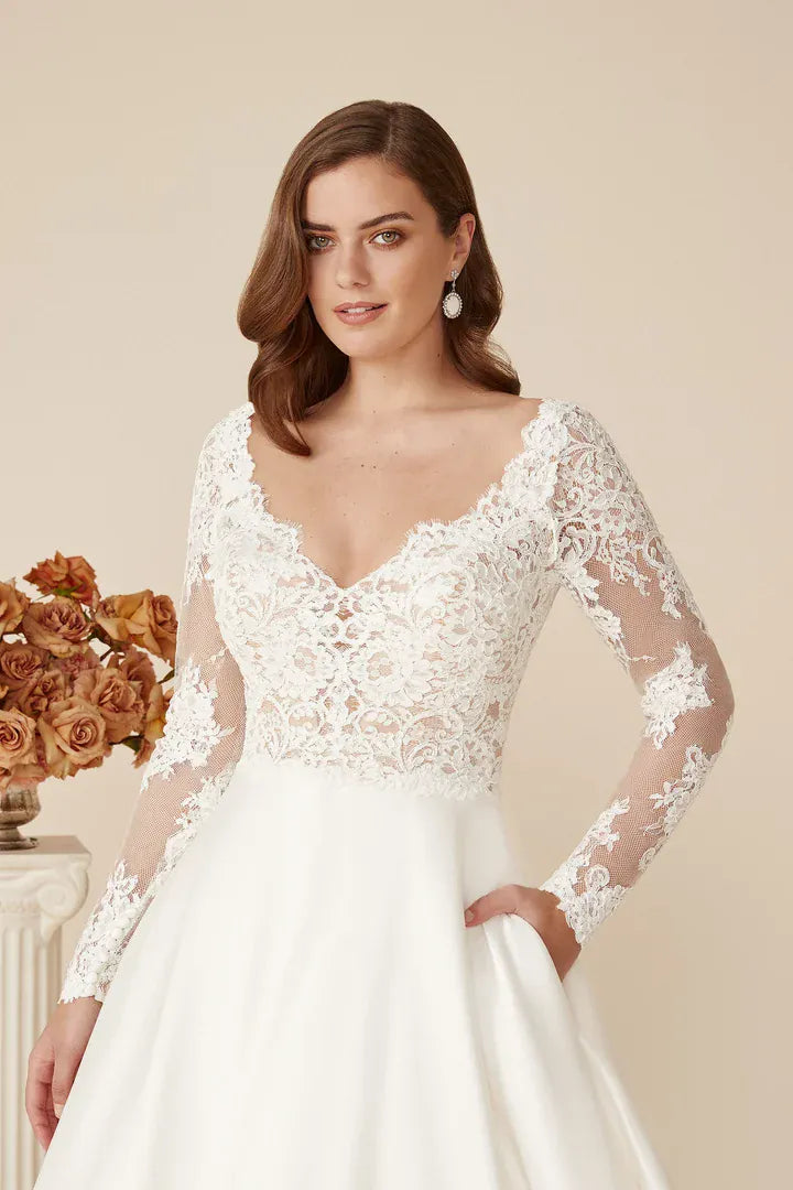 A woman modeling a white wedding dress with a lace bodice, sheer long sleeves, and a full satin skirt against a warm beige studio background.