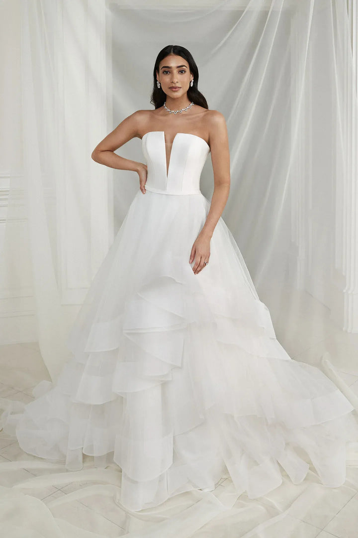 Woman wearing a strapless white wedding dress with a layered skirt against a white curtain background