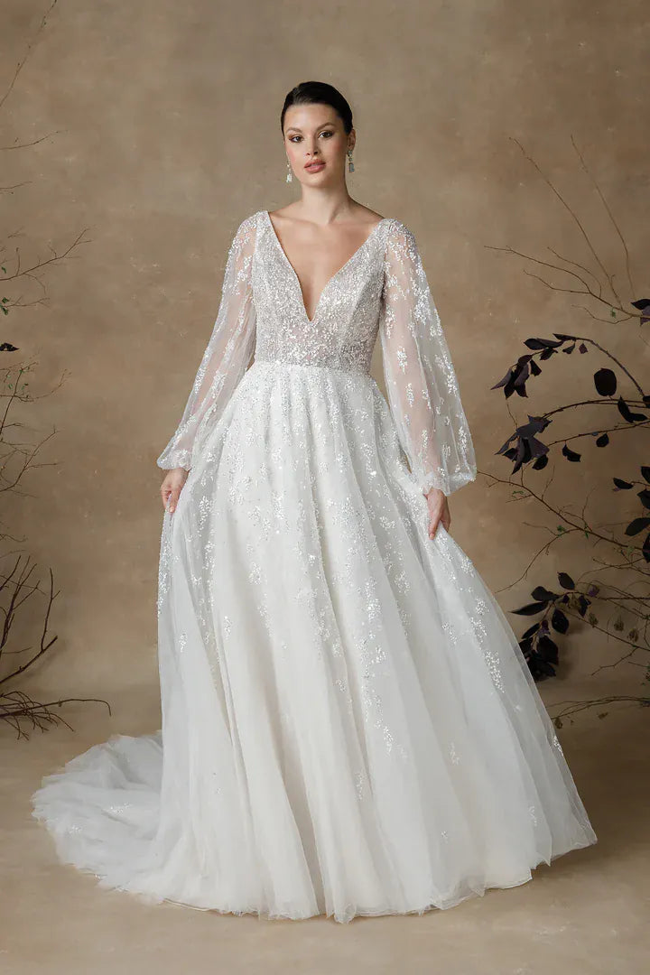 A woman modeling a white bridal gown with sheer long sleeves, a deep V-neckline, and intricate beaded detailing on the bodice and skirt, posed against a warm beige textured backdrop with decorative branches.