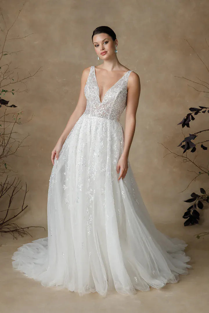 A woman modeling a sleeveless white wedding gown with a deep V-neckline, fitted lace bodice, and full tulle skirt, posed against a warm beige studio backdrop with decorative branches.
