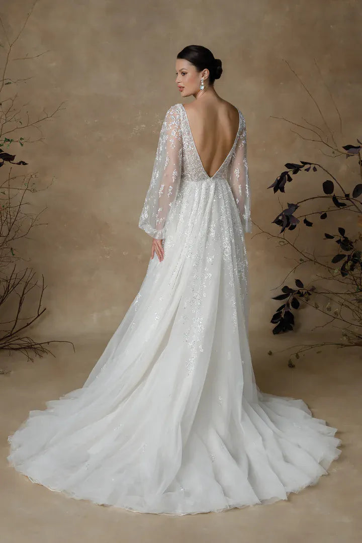 A woman modeling a white bridal gown from the back, showing a deep V back, sheer long sleeves, and a full tulle train, posed against a warm beige studio backdrop with decorative branches.