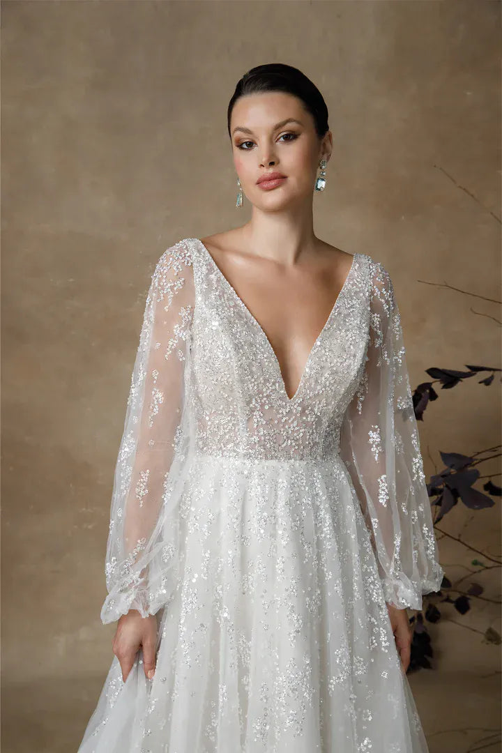 A woman wearing a white lace wedding gown with long sheer sleeves and a deep V-neckline, posed against a warm beige textured backdrop.