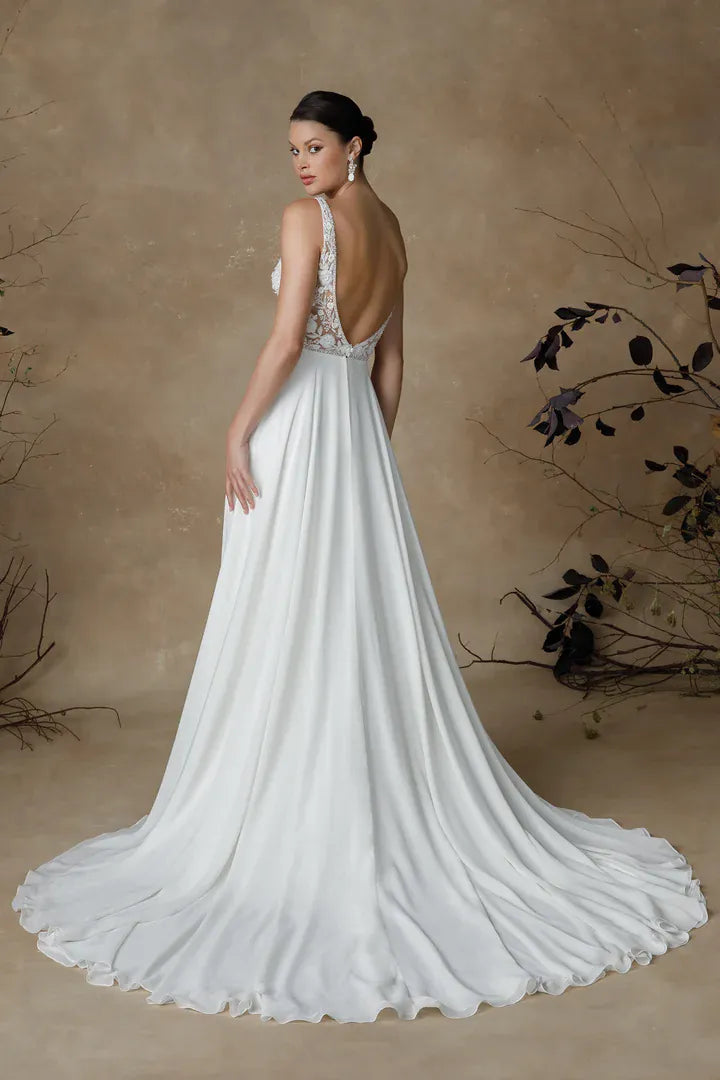 A woman modeling a white bridal gown from the back, showing a fitted bodice with lace detailing and a flowing satin skirt with a long train, set against a warm beige textured backdrop with decorative branches.