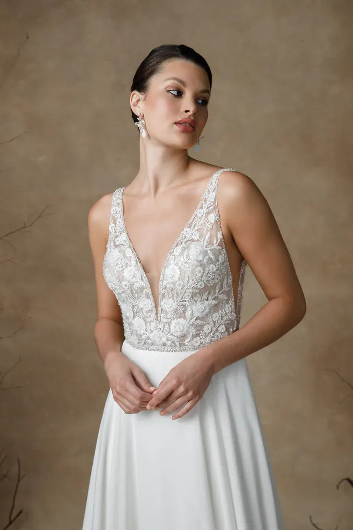 A woman modeling a sleeveless white lace wedding dress with a deep V-neckline and a flowing skirt, posed against a textured warm brown studio backdrop.