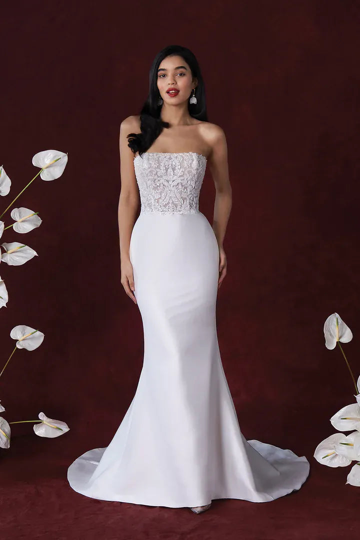 Woman wearing a white strapless gown with lace detailing on a dark background with white flowers.