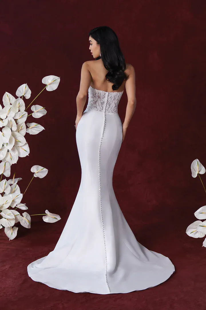 A woman stands with her back to the camera wearing a strapless white mermaid-style wedding gown with a fitted lace bodice, sheer back detail, and a full flared skirt on a dark burgundy backdrop with white floral props.