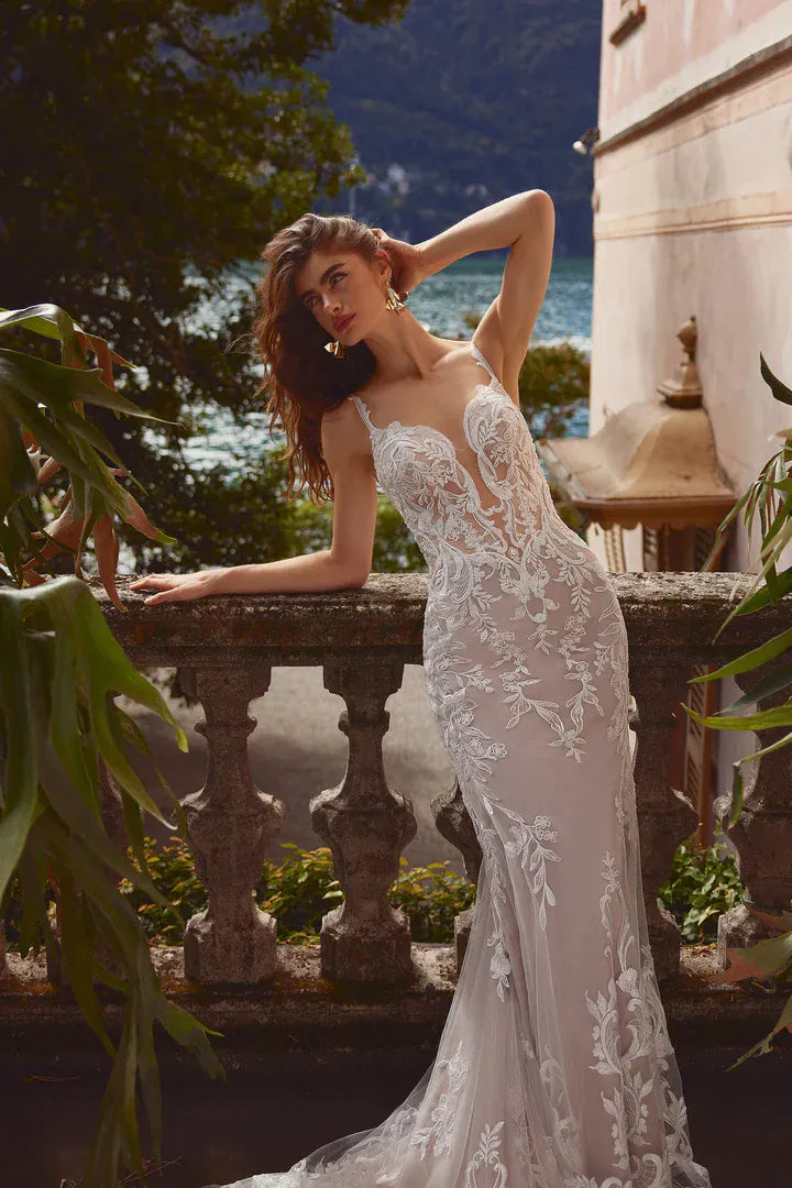 A woman wearing a fitted white lace wedding gown with floral embroidery leans against an outdoor stone balustrade, with water, trees, and a building visible in the background.