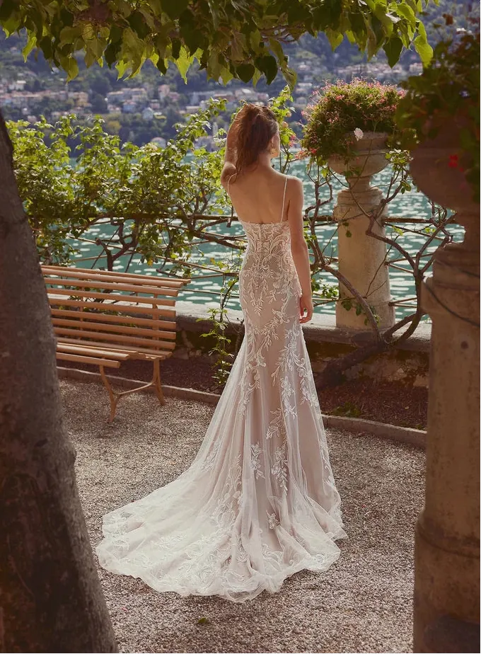 Woman in a lace wedding dress standing by a scenic lakeside with mountains in the background.