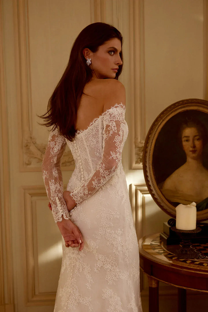 Woman in a lace wedding dress standing in a room with a painting on the wall.