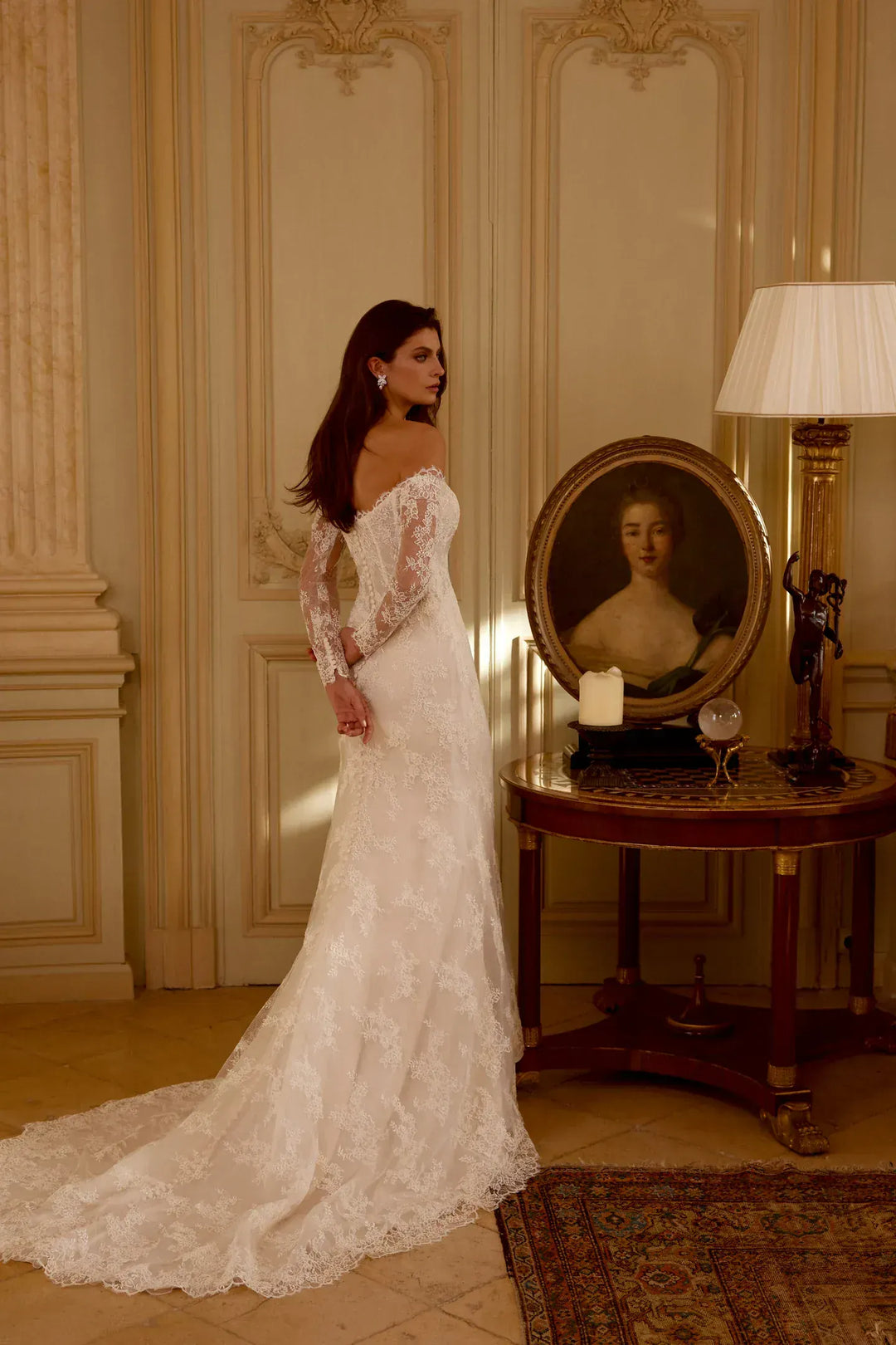 Woman in a white lace dress standing in an elegant room with a portrait and lamp.