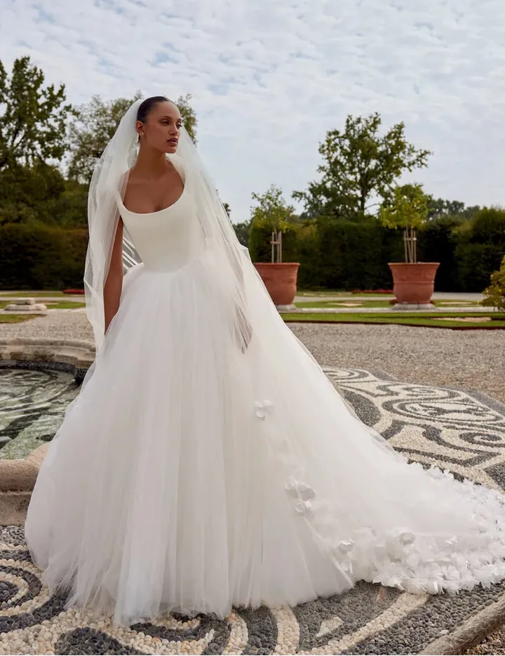Bride in a white wedding dress with a long veil standing in an outdoor setting with trees and a fountain.