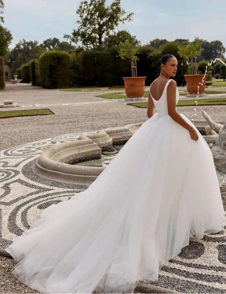 Woman in a white wedding dress standing in an outdoor setting with greenery and a fountain.