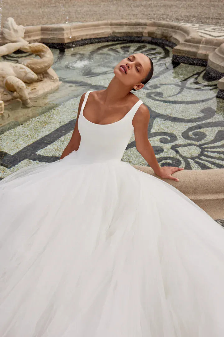 A woman wearing a white wedding dress with a fitted bodice and a voluminous tulle skirt, leaning against a stone fountain in an outdoor setting.