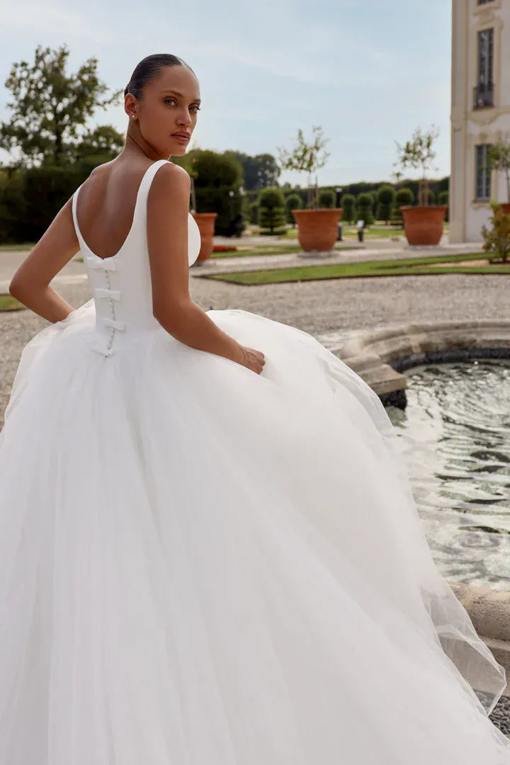 A woman wearing a white wedding gown stands outdoors beside a fountain in a formal garden, photographed from the back to show the lace-up back and full tulle skirt.