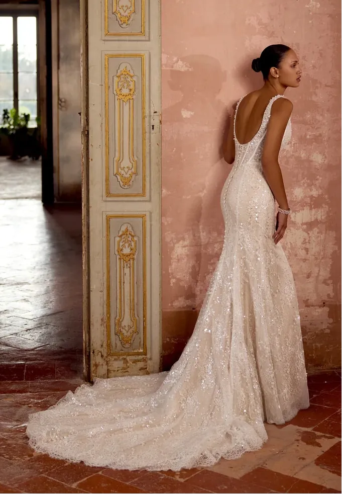 A woman wearing a fitted ivory lace wedding gown with a long train stands in profile against a pink plaster wall in an indoor setting with an ornate door and tiled floor.