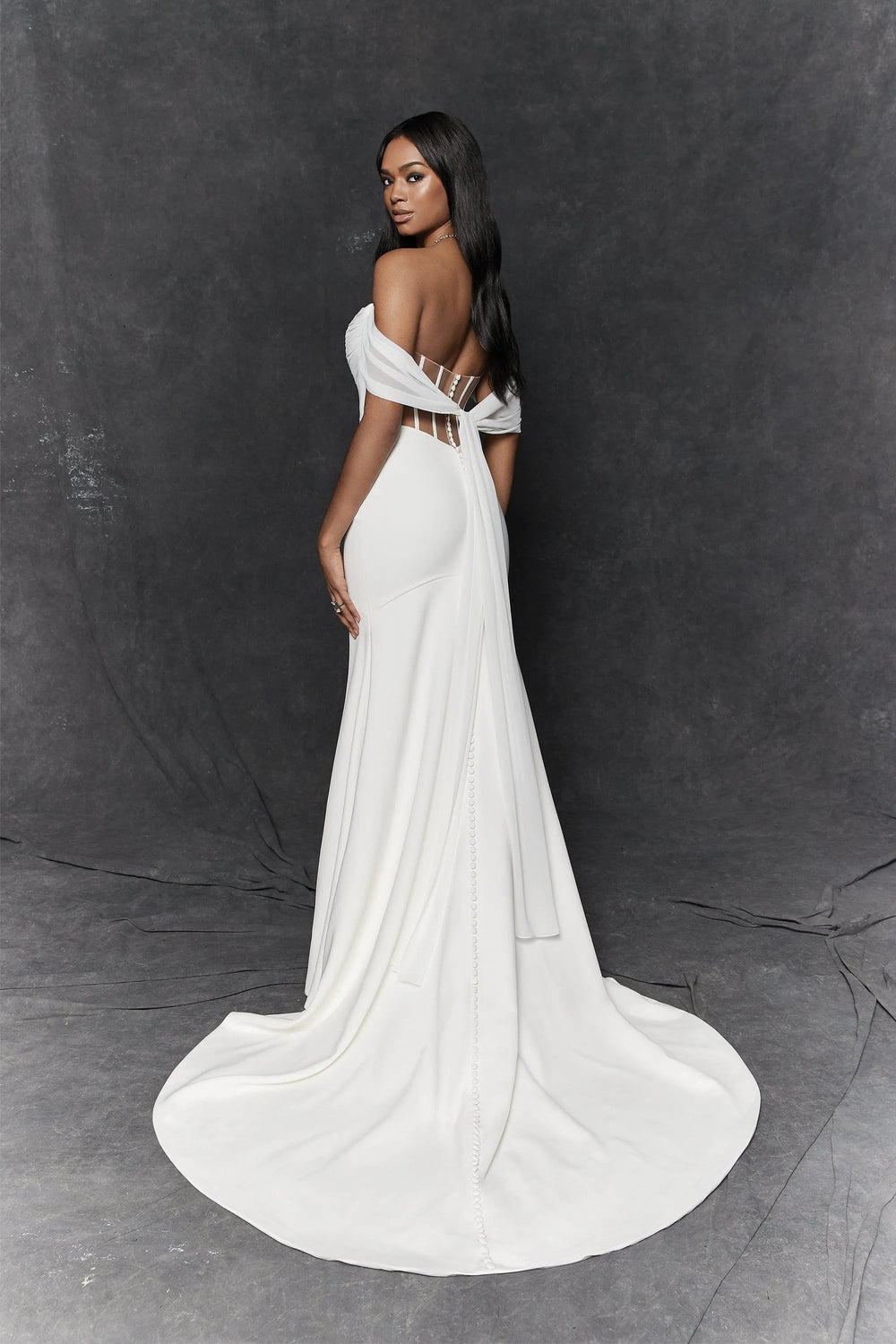 A woman modeling a floor-length white gown with an off-the-shoulder design, sheer side panels, and a dramatic train, photographed against a dark gray studio backdrop.