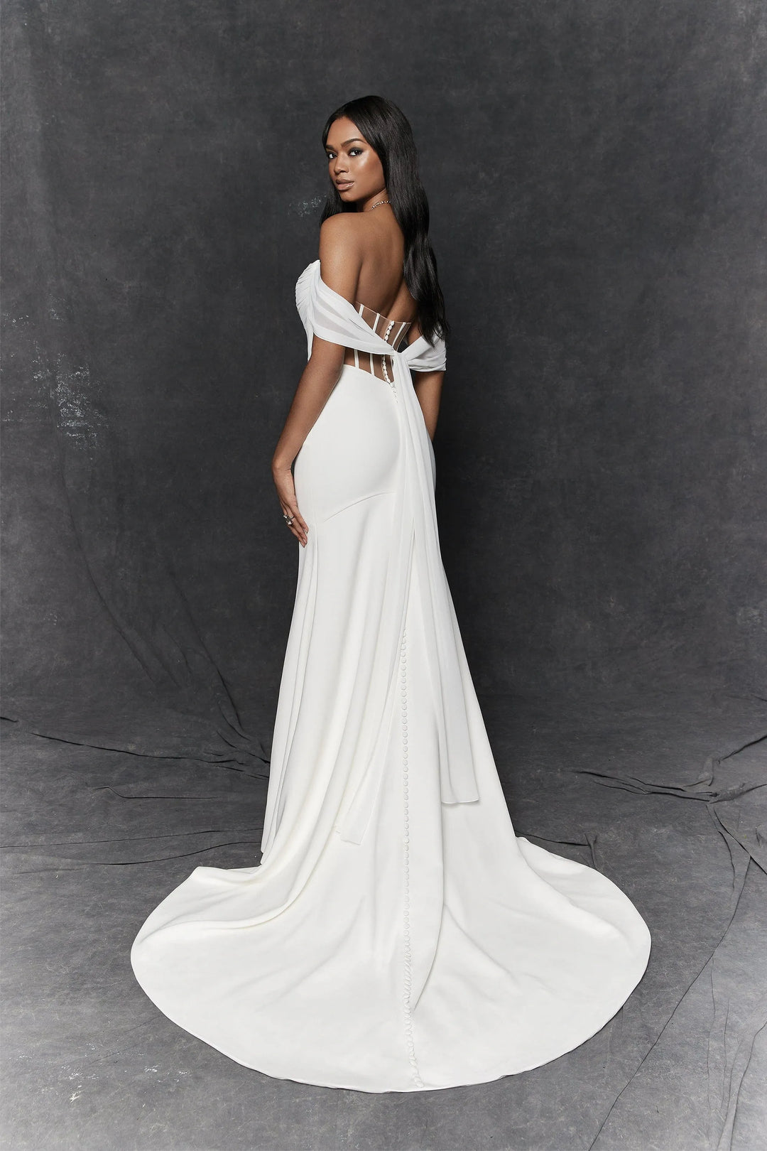 A woman modeling a floor-length white gown with an off-the-shoulder design, sheer side panels, and a dramatic train, photographed against a dark gray studio backdrop.