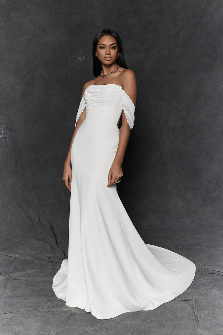 A woman modeling an off-the-shoulder white mermaid-style gown with a fitted bodice and flowing skirt, standing against a dark gray studio backdrop.