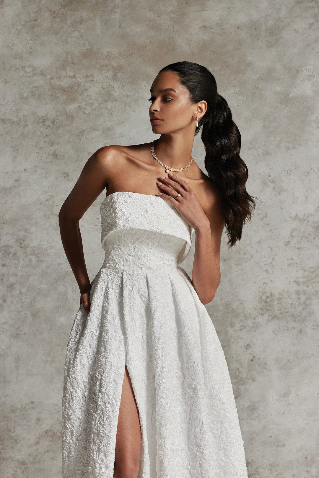 Woman wearing a strapless white wedding dress with a high slit on a textured gray background