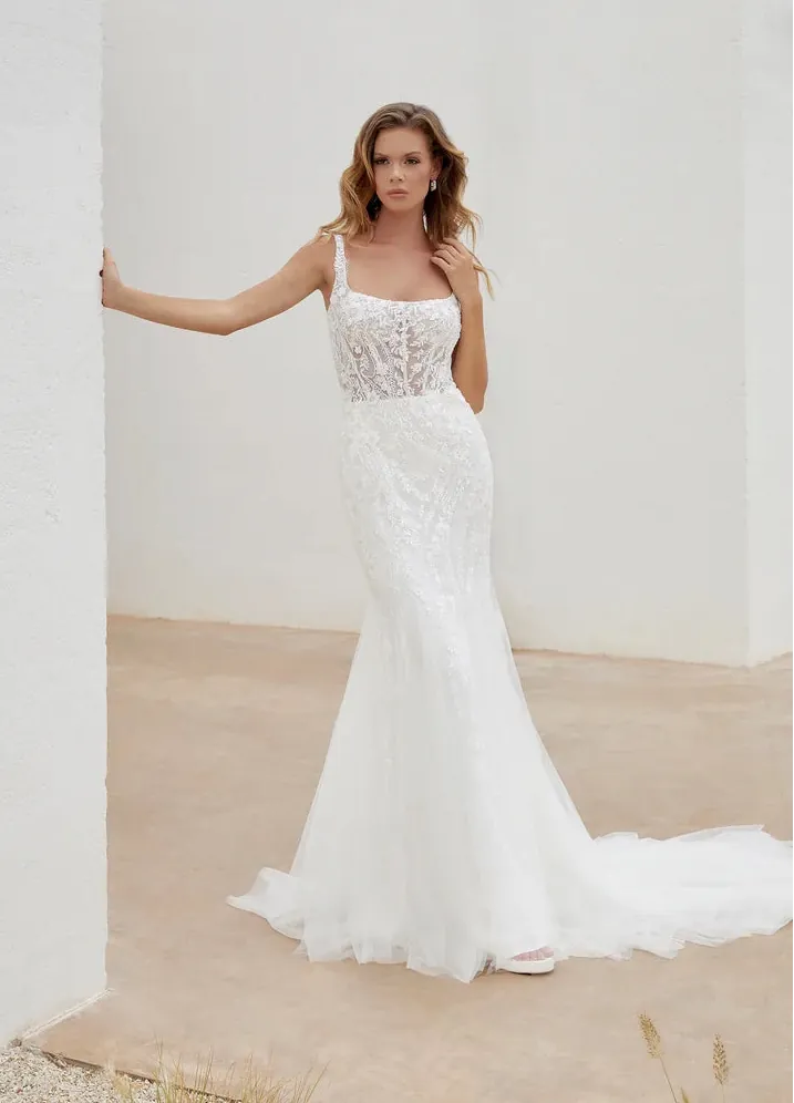 A woman modeling a white lace mermaid-style wedding gown with thin straps and a long sheer tulle train, standing in a bright white architectural setting with a beige floor.