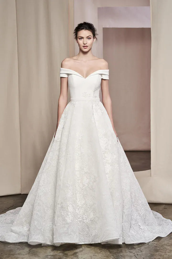 An off-the-shoulder white lace wedding gown with a sweetheart neckline and a full ball gown skirt, shown on a model in a studio setting.