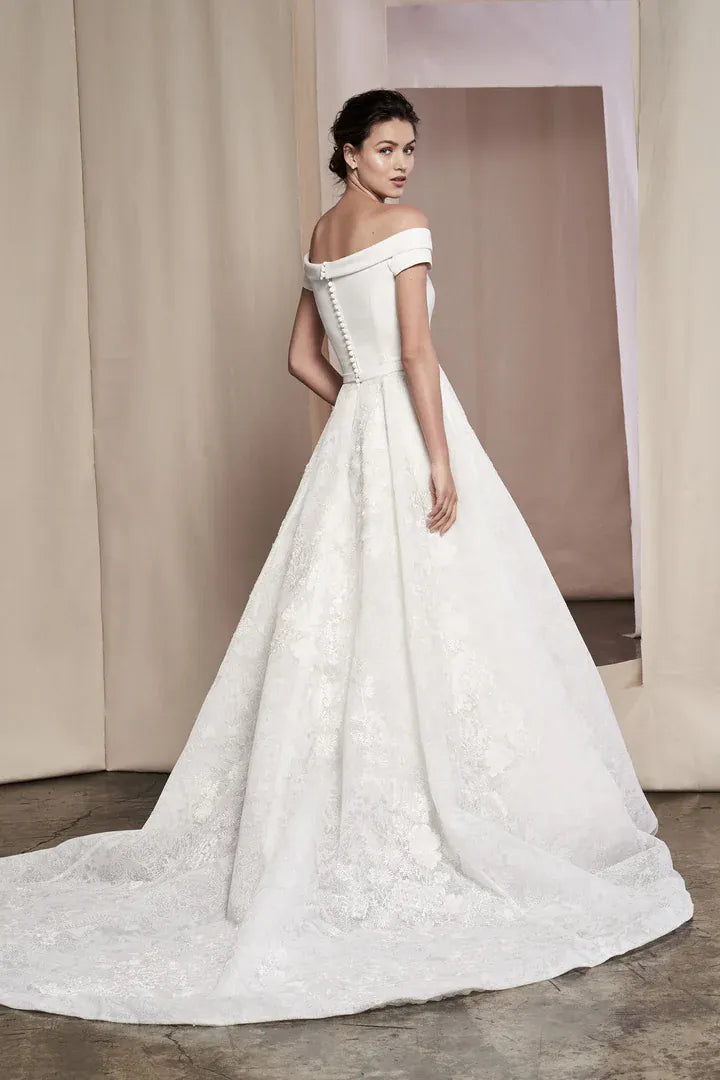 A woman modeling an off-the-shoulder white ball gown with a structured lace bodice, fitted waist, and full skirt with a long train in a softly lit studio setting.