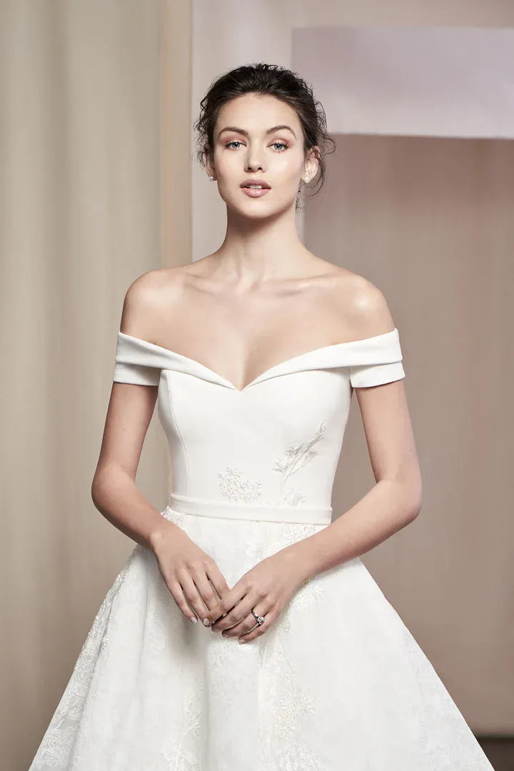 A woman modeling an off-the-shoulder white wedding gown with a sweetheart neckline, fitted bodice, and full skirt against a soft beige studio backdrop.