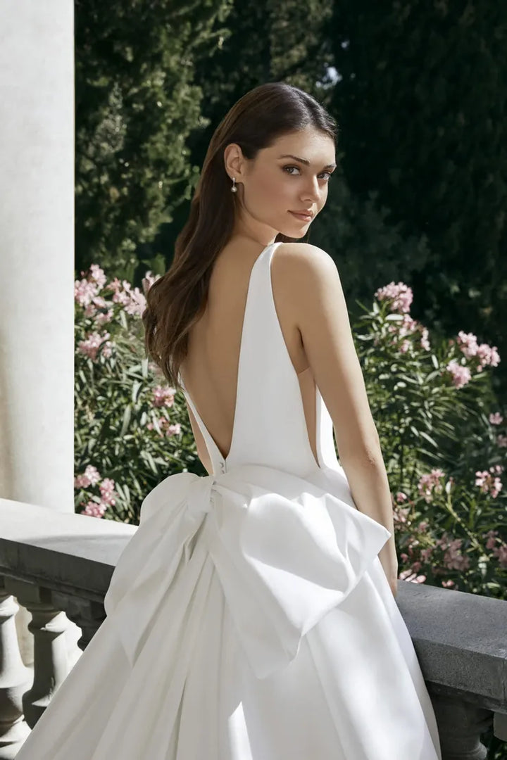 Woman in a white wedding dress with a large bow standing outdoors.