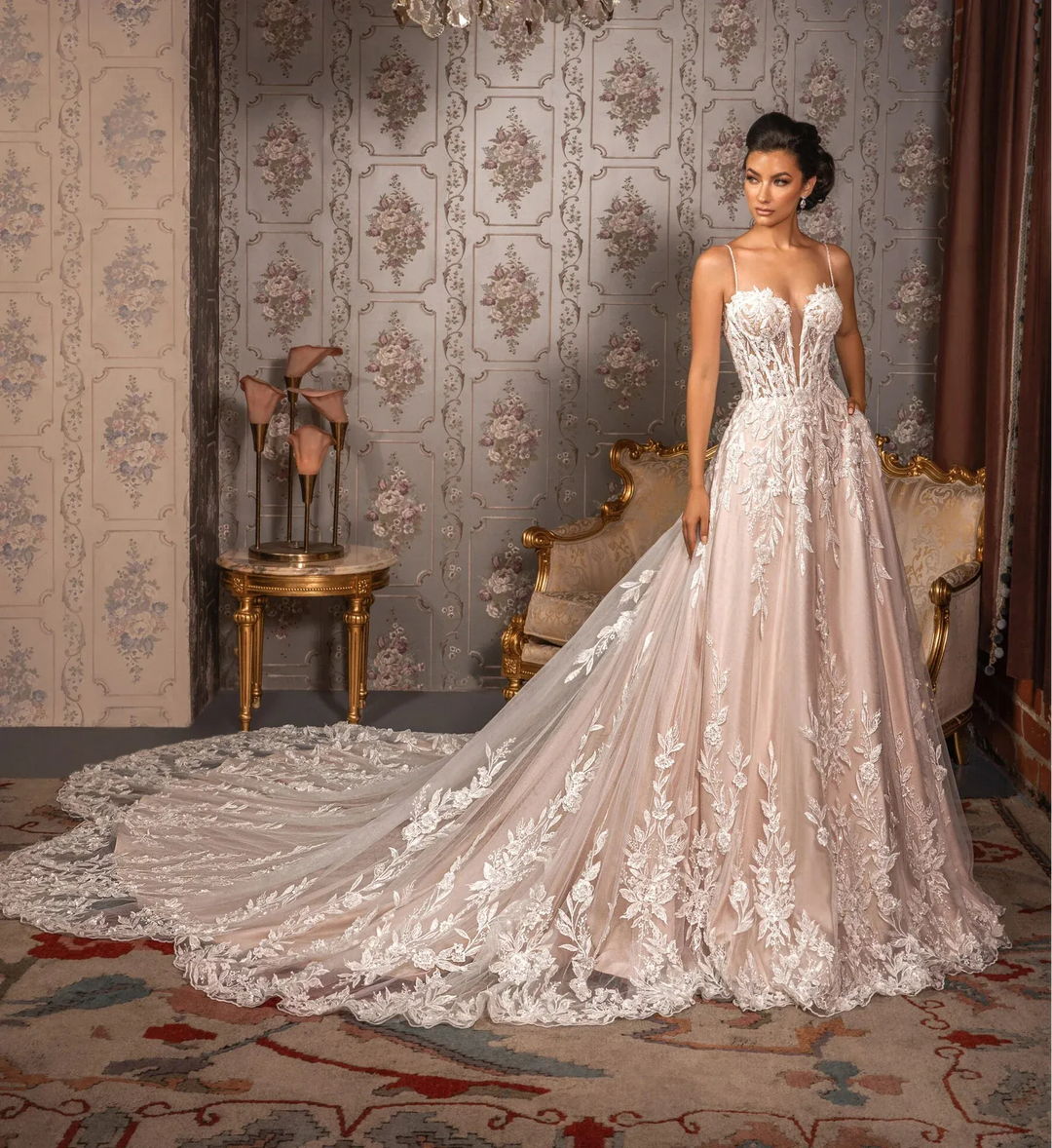 Woman in a white lace wedding dress standing in an ornate room with a chandelier.