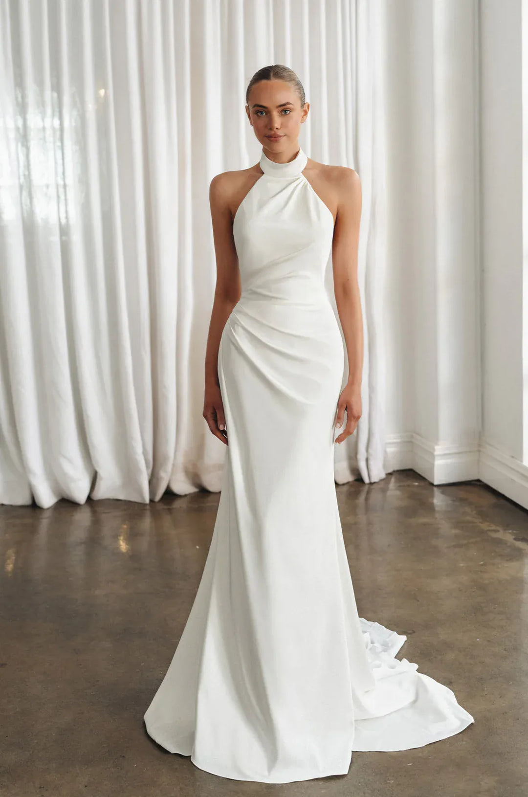 A woman modeling a floor-length white halter gown with a draped, gathered bodice and a slight train, standing in a bright room with white curtains and a polished concrete floor.