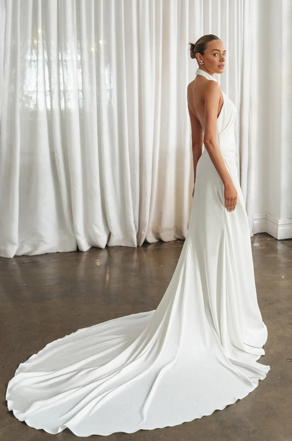 Woman in a white wedding dress with a long train against a white curtain backdrop, featuring the brand 'Osquernovia'.