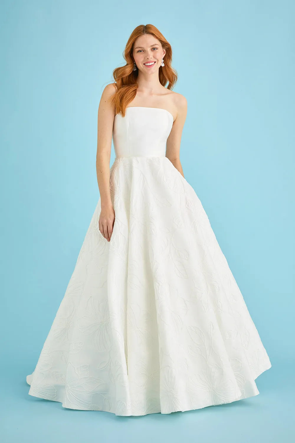 A woman modeling a strapless white ball gown with a structured bodice and a full, flowing skirt against a solid light blue studio background.