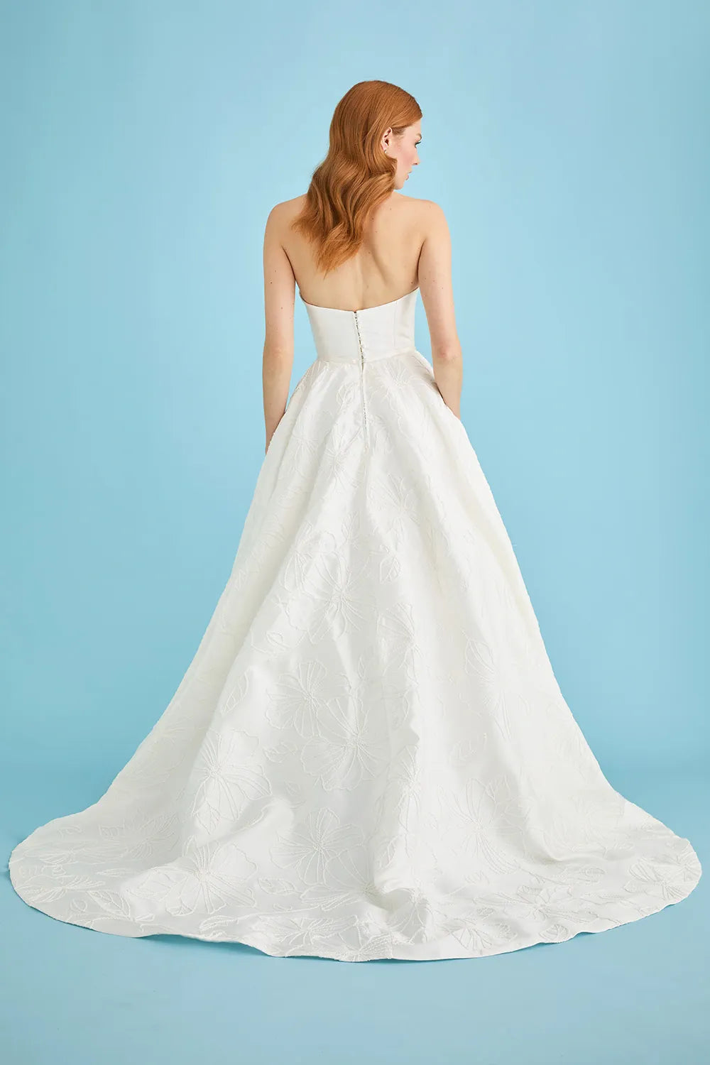 A woman with red hair is shown from the back wearing a strapless white wedding gown with a structured bodice and a full, flowing skirt on a light blue studio background.