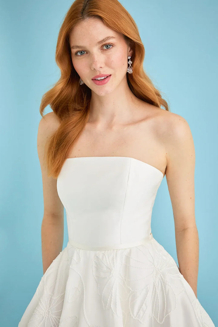 A woman with long red hair wearing a strapless white wedding dress with a structured sweetheart bodice and a full textured skirt, posed against a solid light blue background.