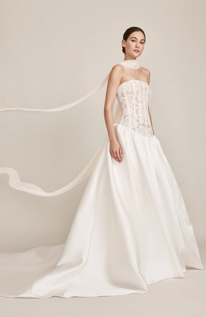 A woman modeling a strapless white wedding gown with a high neck, lace bodice, and full satin skirt with a long train against a soft beige studio background.