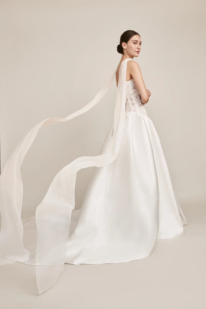 A woman modeling a white bridal gown from the back, showing a sheer long veil and a structured satin skirt, against a plain warm beige studio background.