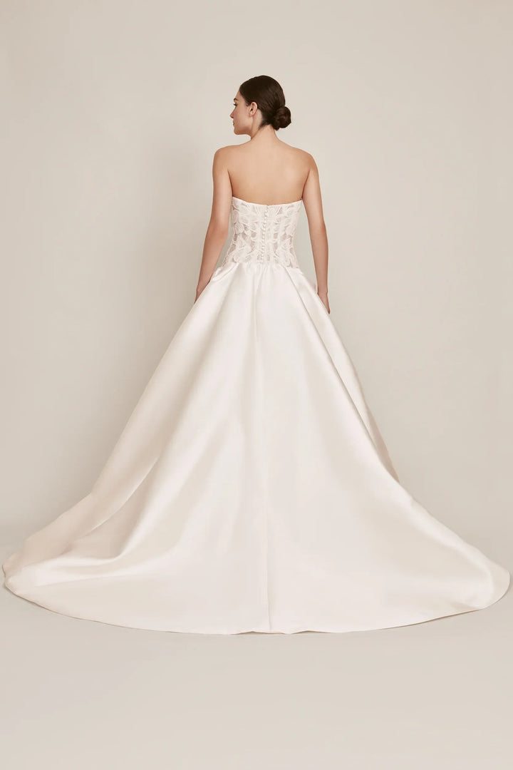 A woman modeling a strapless white wedding gown from the back, showing a structured lace corset-style bodice and a full satin train against a plain warm beige studio background.