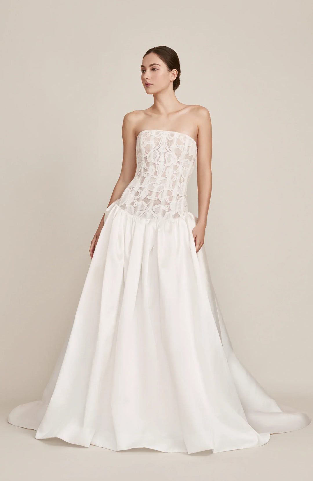 A woman modeling a strapless white wedding gown with a lace corset-style bodice and a full satin skirt, photographed against a plain warm beige studio background.