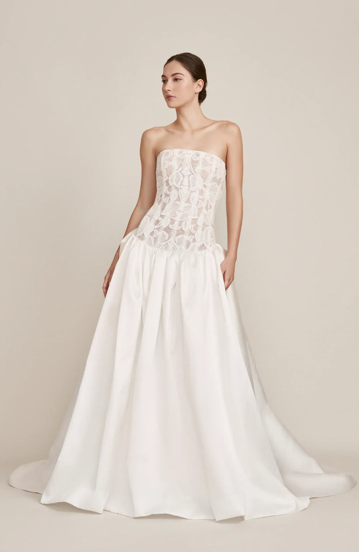 A woman modeling a strapless white wedding gown with a lace corset-style bodice and a full satin skirt, photographed against a plain warm beige studio background.